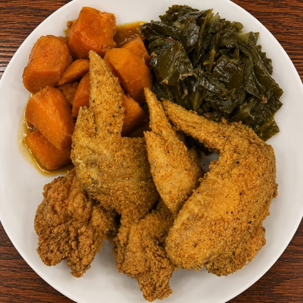 A plate of fried chicken wings, candied yams, and collard greens on a wooden table.