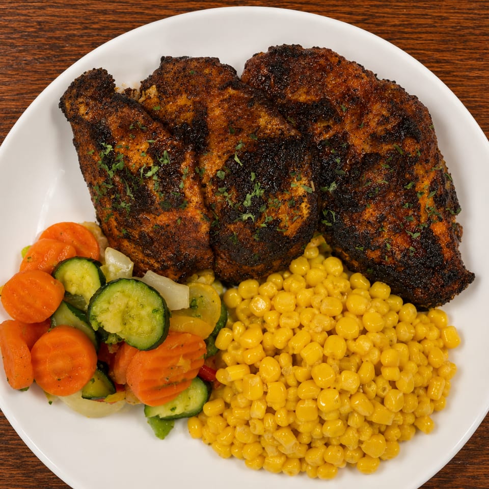 Three blackened chicken breasts with a side of mixed vegetables and corn on a white plate.