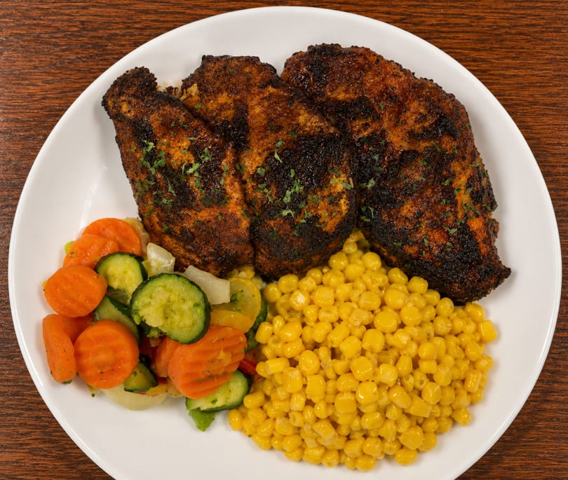 Three blackened chicken breasts with a side of mixed vegetables and corn on a white plate.